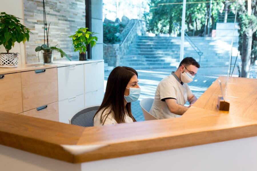 A woman with long dark hair and a man wearing glasses work behind a wooden reception desk in a well-lit office. Both are wearing face masks. The setting includes modern cabinets, potted plants, and a large window revealing trees and a stone stairway in the background